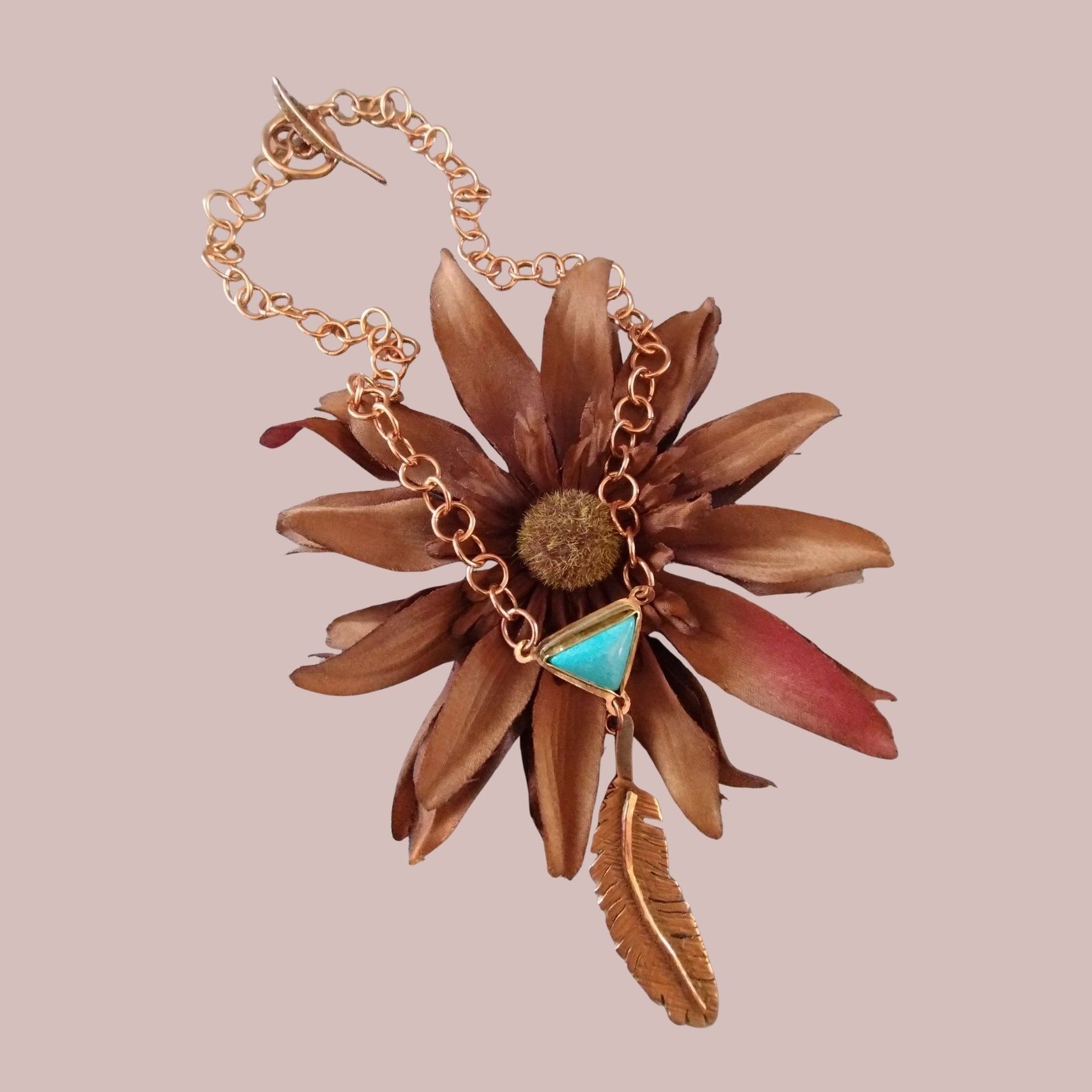 Handmade custom feather necklace with turquoise cabochon displayed on a brown flower, hand crafted by SoufiiArte Studio.